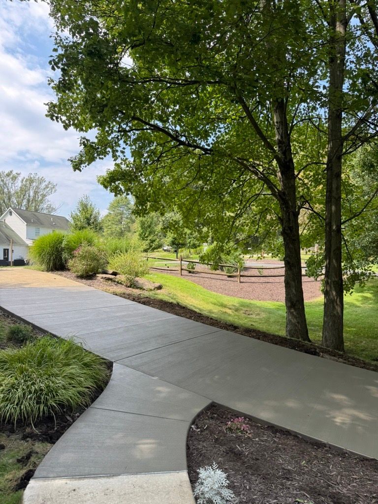 Curved concrete walkway through a landscaped suburban yard with trees and a house on the left.