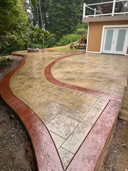 Curved stamped concrete patio with red border beside a house and wooded yard
