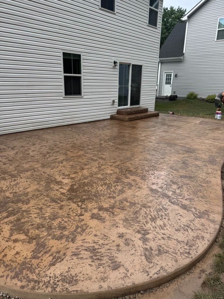 Stamped concrete patio beside a white house, wet from rain, with a curved edge and back door steps