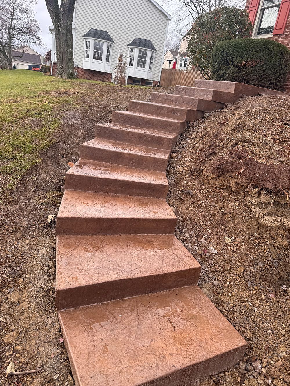 A curved outdoor staircase made of reddish-brown concrete leading up a grassy hill toward a house.