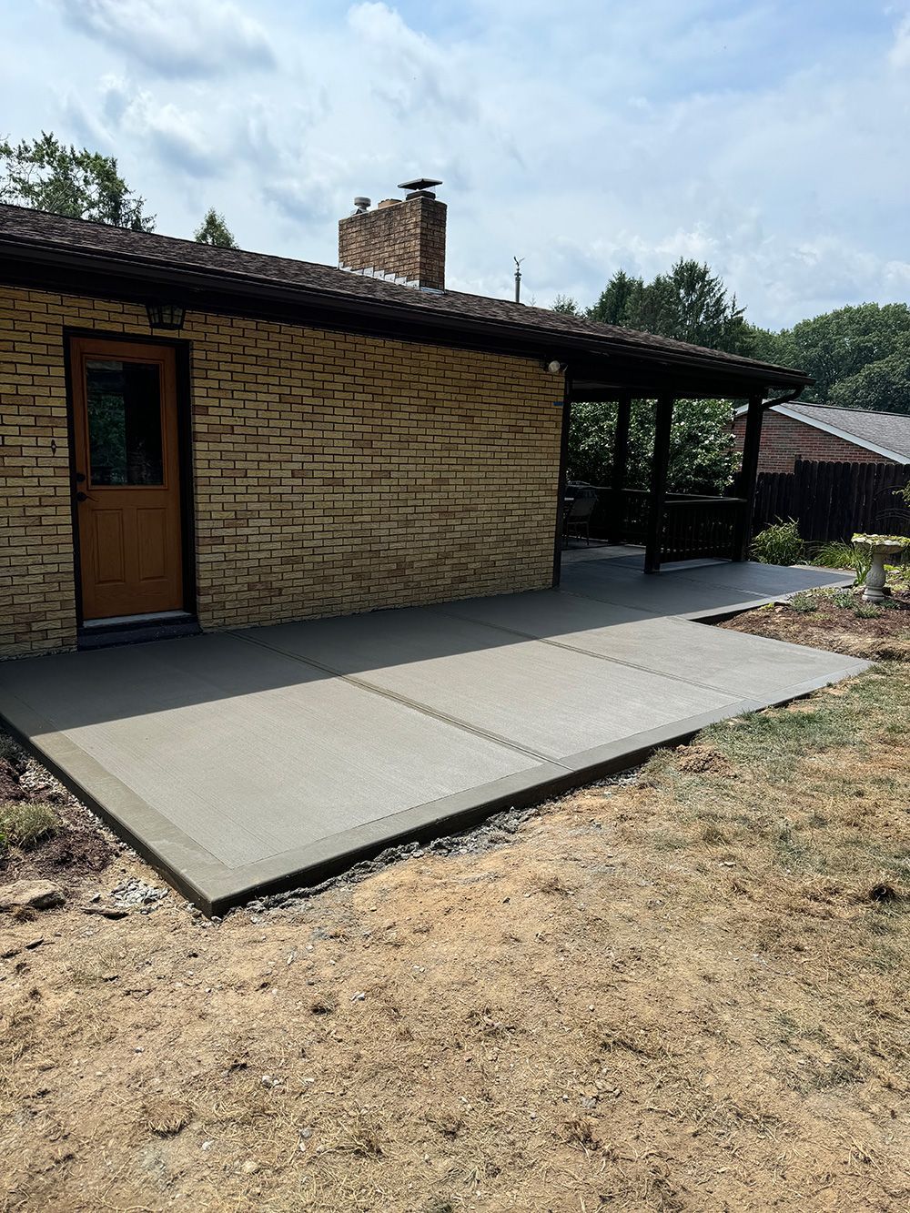 Newly poured gray concrete patio extending from a yellow brick house with a brown door and covered porch.