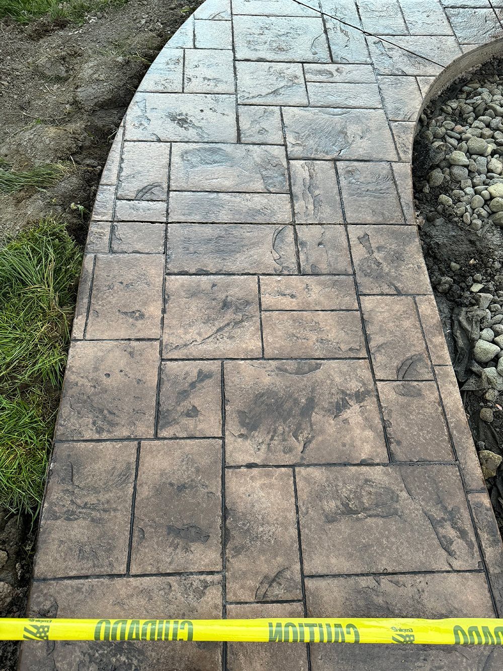 A curved walkway made of rectangular stamped concrete pavers in shades of brown and gray, edged with gravel and grass.