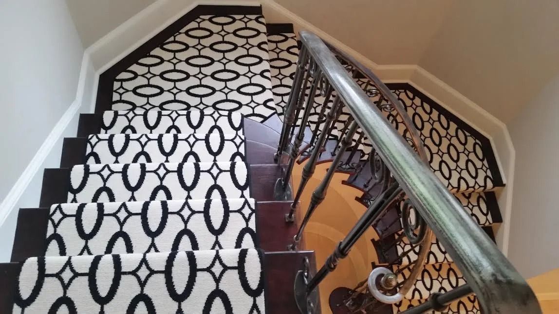 A high-angle view of a staircase featuring a white runner carpet with a black geometric oval pattern and a metal railing.