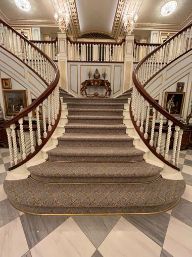 A grand, ornate foyer featuring a sweeping double staircase with white railings, carpeted steps, and marble flooring.