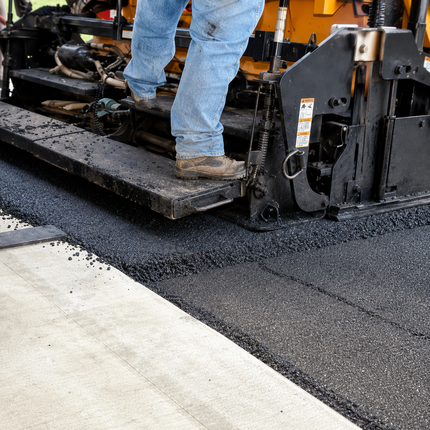 Worker standing on asphalt paver laying fresh black pavement beside a concrete curb