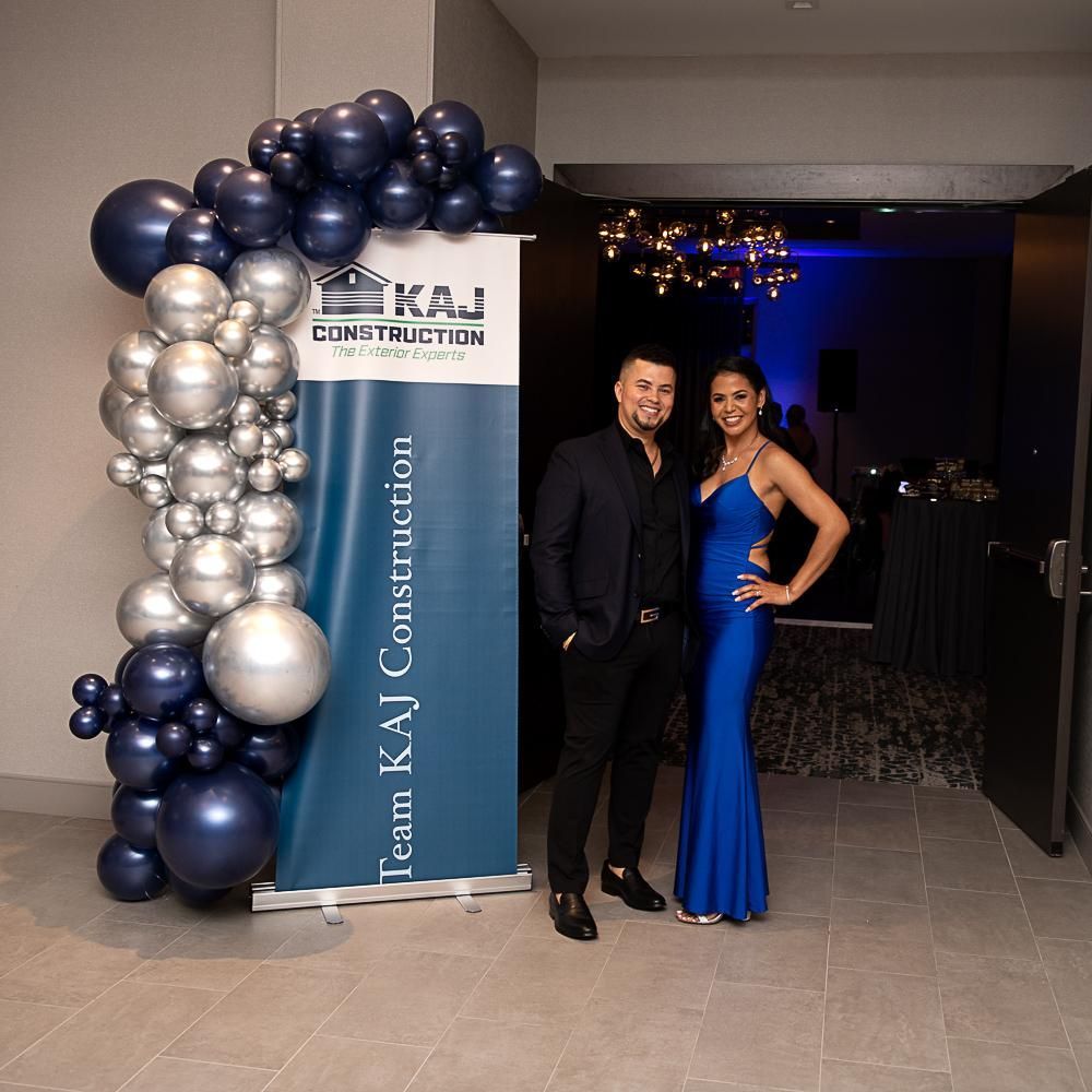 A man and a woman are standing in front of a sign that says Team KAJ Construction.
