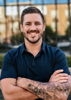 Man with crossed arms, smiling, wearing a navy polo shirt with tattoos on his arm, outdoors.