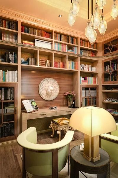 Cozy home library with built-in bookshelves, a desk, and seating, illuminated by a chandelier and lamps.