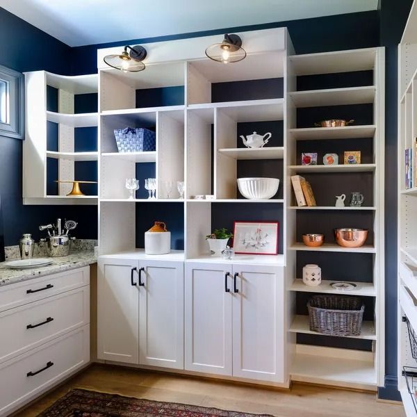 White pantry with dark blue accent wall, shelves displaying various items, white cabinets, and two sconce lights.