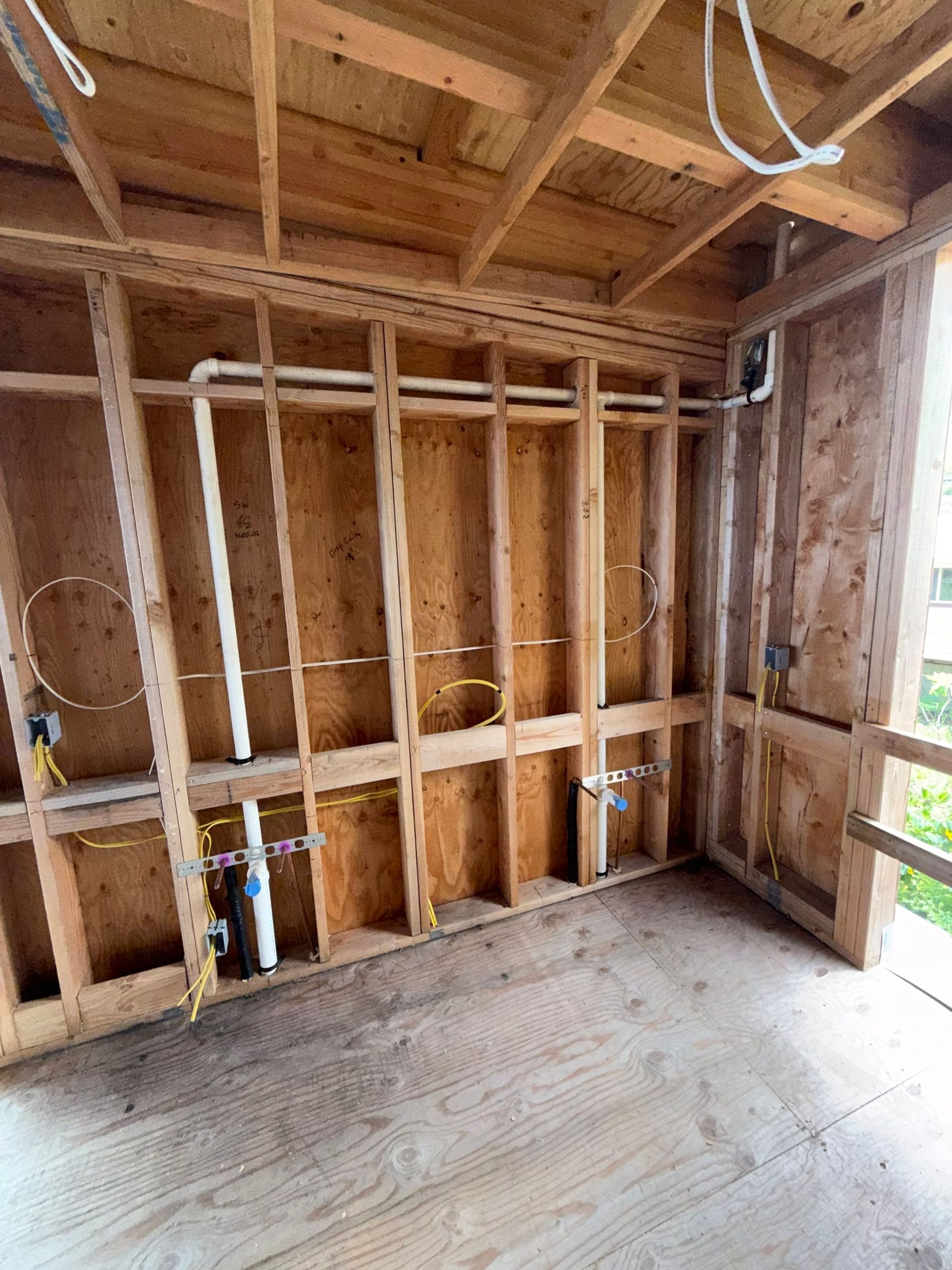 Unfinished interior wall framing with exposed wooden studs, plywood floor, and open window light.