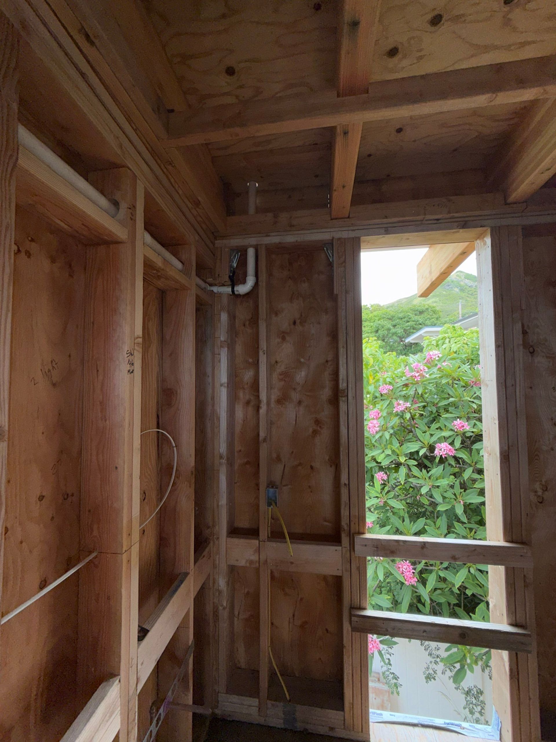 Wooden interior wall framing with a window opening to green foliage and pink flowers outside