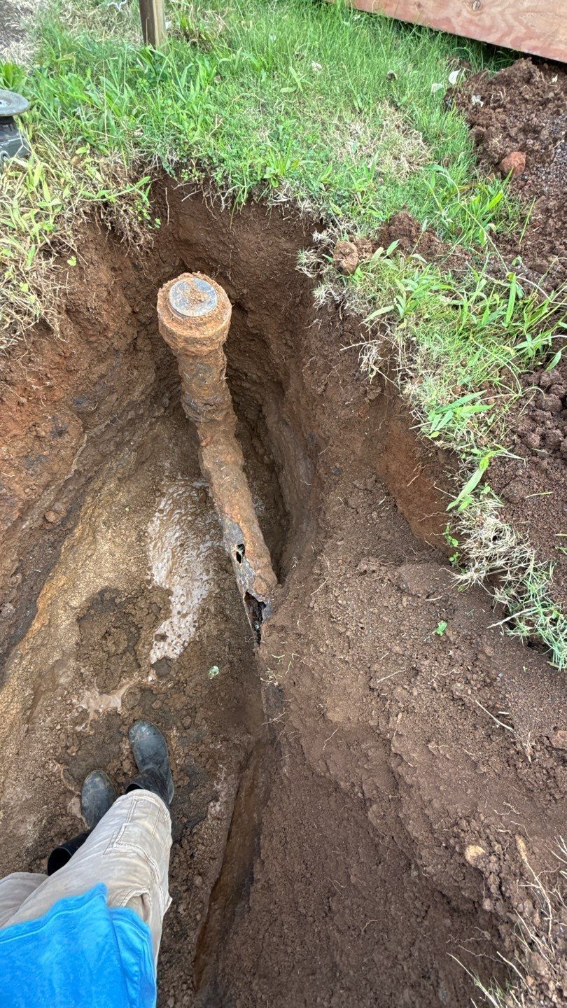 Open trench in soil with a rusted pipe; a person’s boot is visible at the edge.