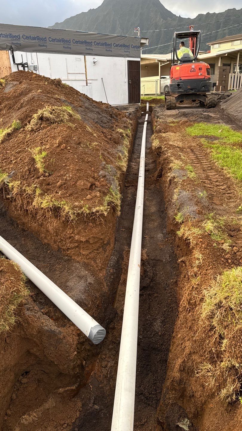 Excavator digging a trench with large white pipes laid along the muddy ground near buildings and hills