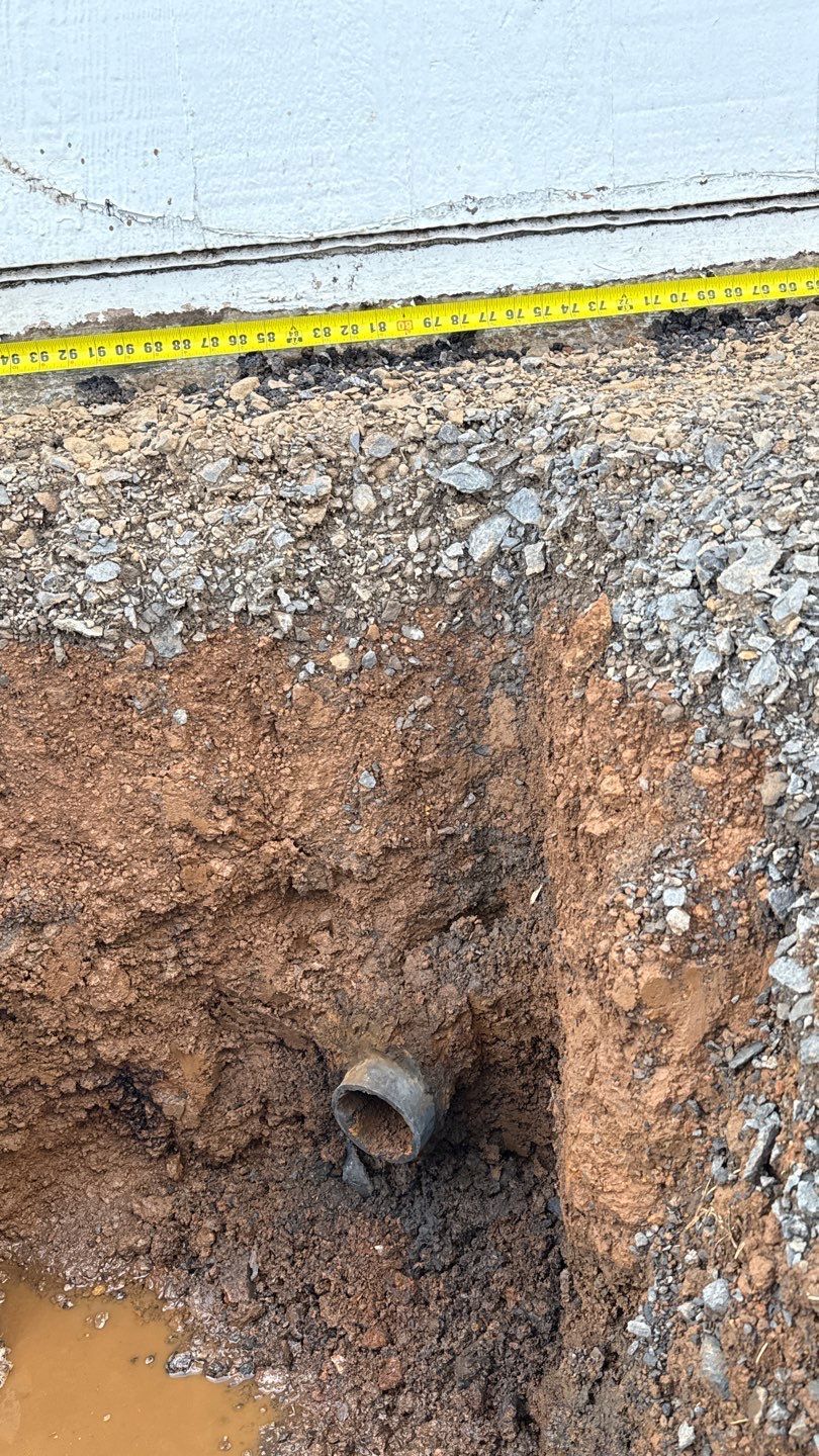 Excavation trench with exposed pipe opening in brown soil beside a gravel roadside.