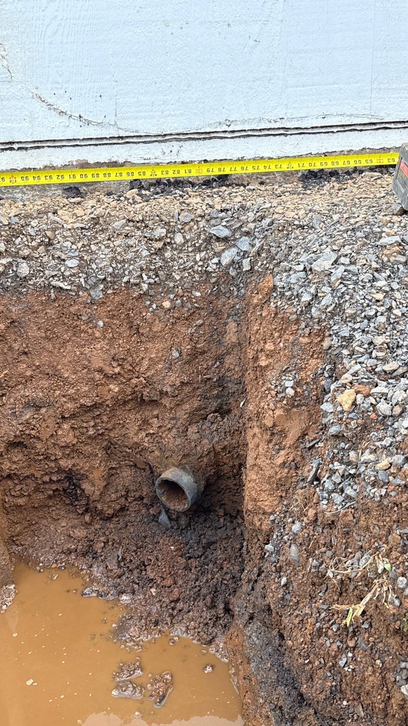 Excavated trench with a pipe opening in muddy soil beside gravel and a wall or curb edge