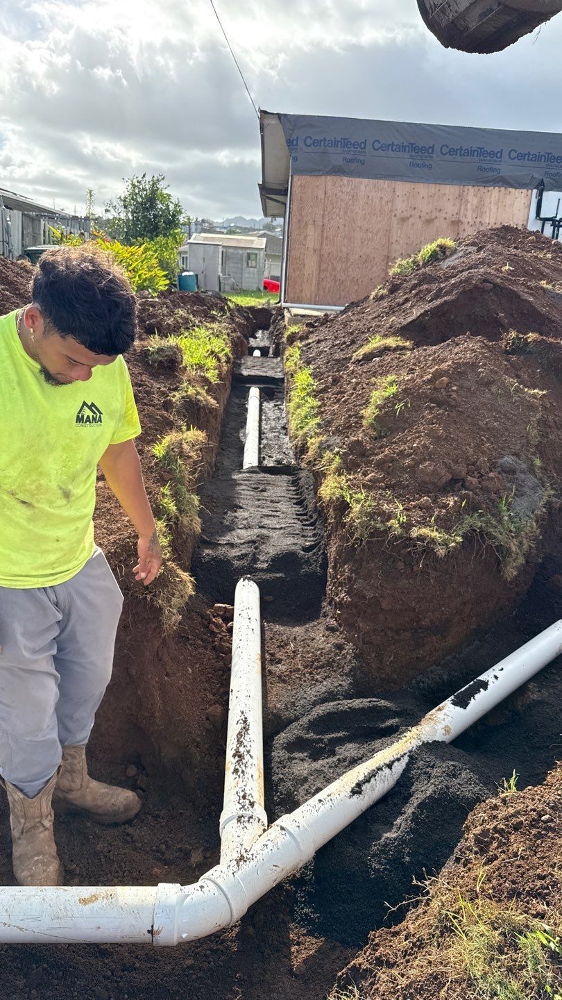 Worker in neon shirt standing by a trench with white pipes beside a large dirt mound outdoors