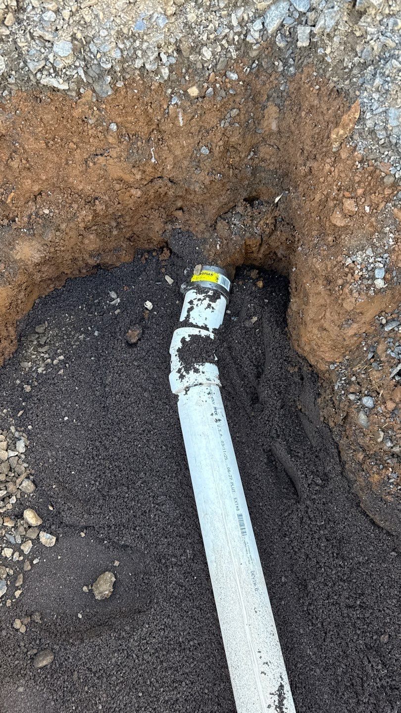 White pipe with elbow joint partially buried in a dirt trench, surrounded by gravel and soil.