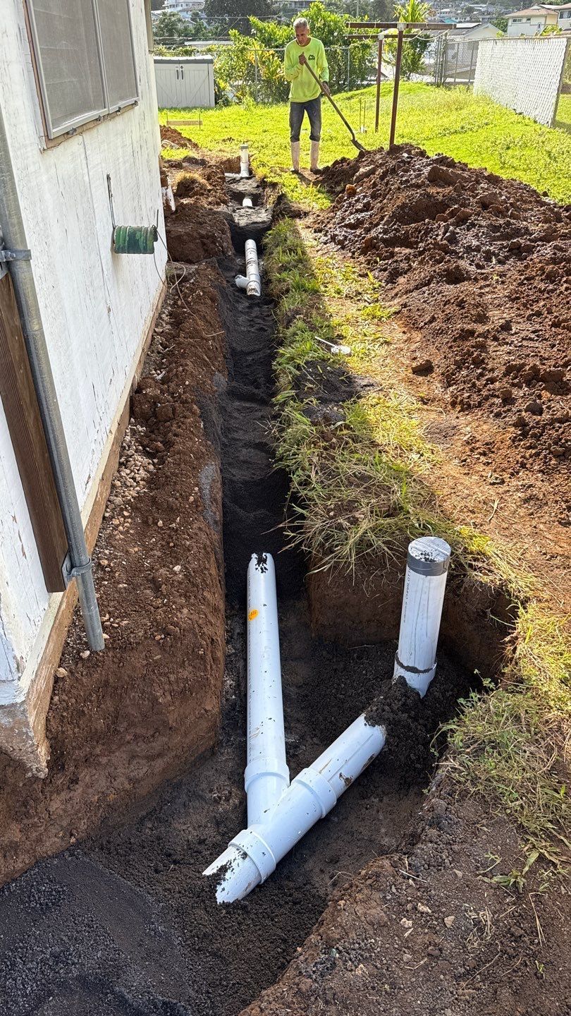 Trenched yard with white PVC pipes and a worker standing near a house on a dirt slope