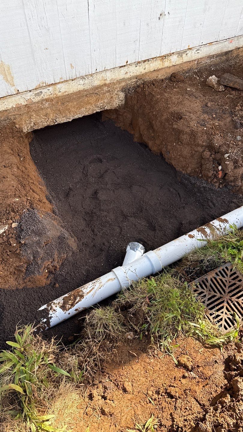 Large hole beside a building wall with exposed soil, a white pipe, and a drain grate nearby