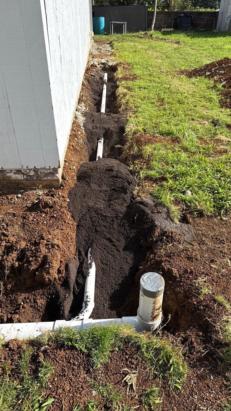 A narrow trench beside a house reveals exposed pipes and fresh soil in a grassy yard.