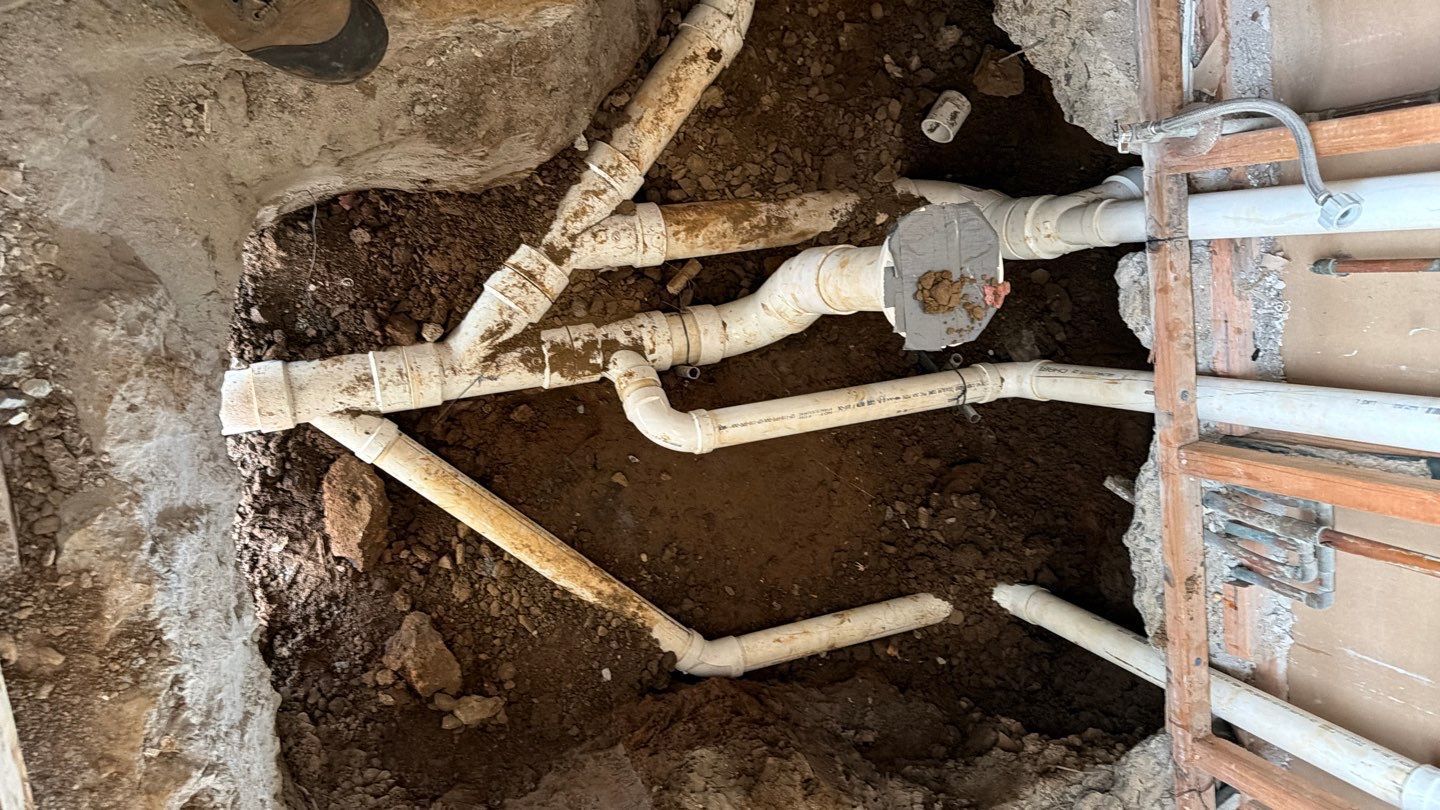 Exposed plumbing pipes and fittings in a dirt trench beside a wall