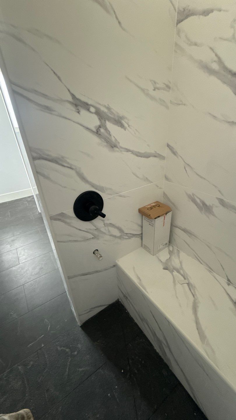 Bathroom wall with marble tile, a round wall opening, and a small wooden shelf above a black floor.