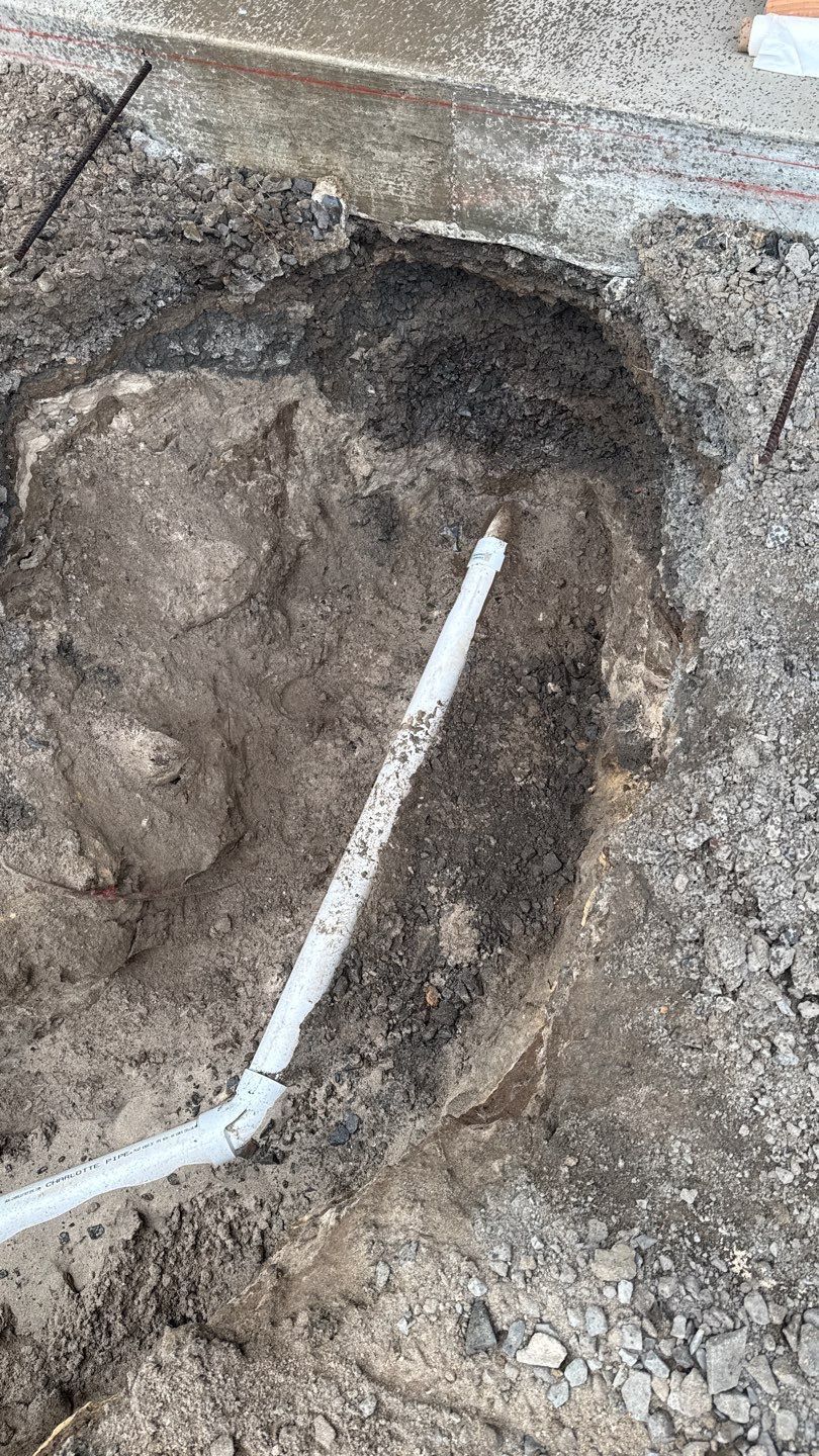 Excavated hole in dirt with a white pipe or conduit running through it near a concrete edge