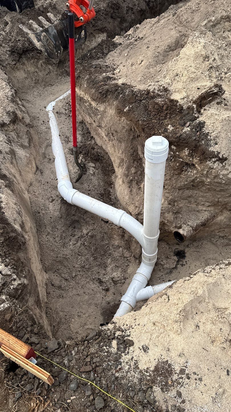 White PVC plumbing pipes installed in a dirt trench, with a red marker stake beside them.