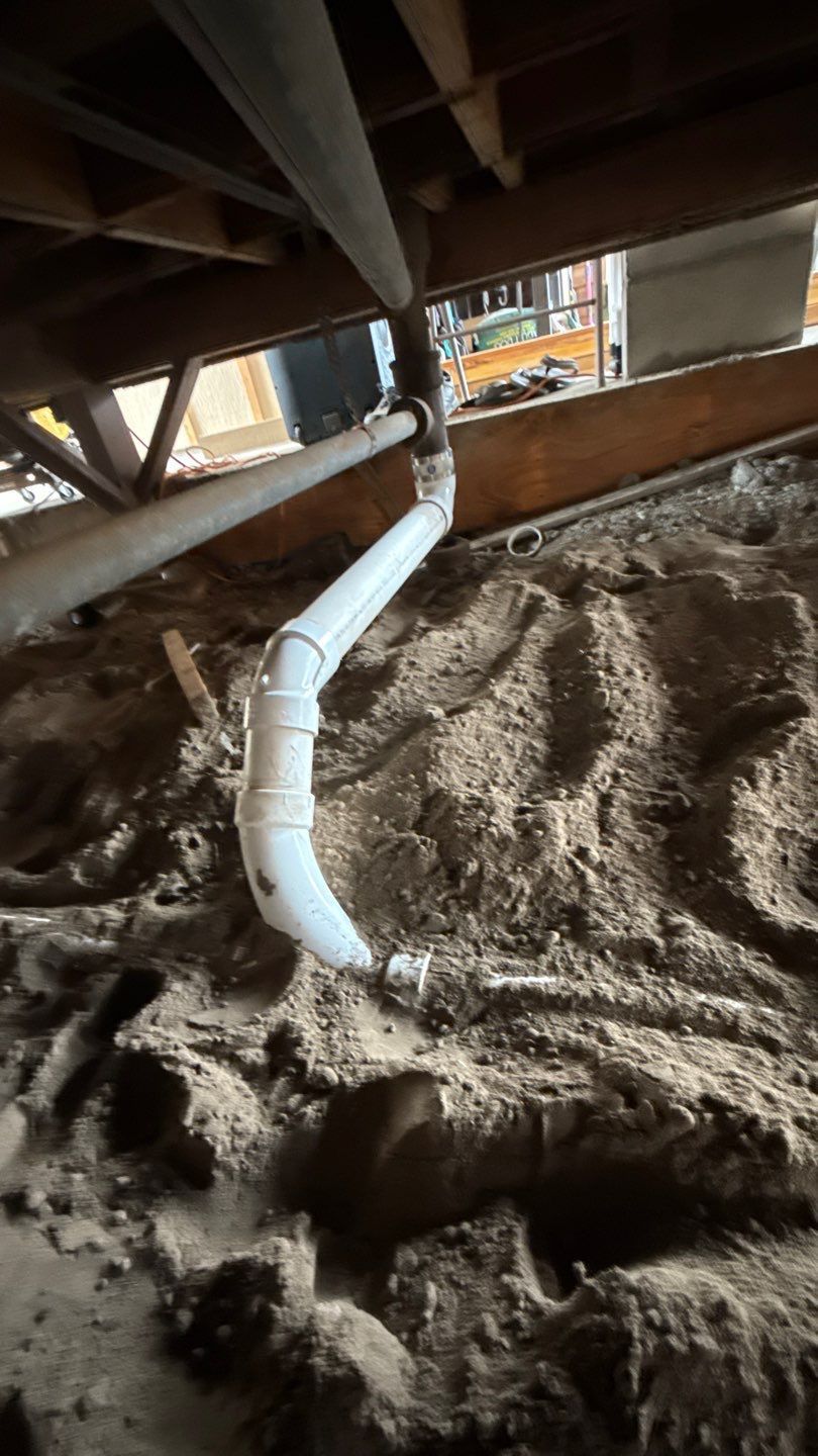 White PVC pipe runs through a dirt crawlspace beneath floor joists.