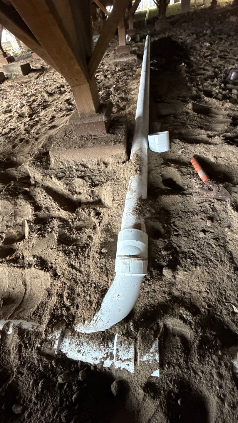 White PVC pipe and fittings running along dirt under a wooden structure, with a small gray box nearby.