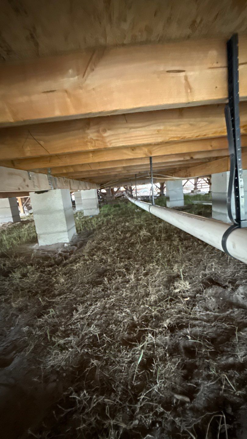 Crawlspace under a house with wooden beams, concrete piers, exposed pipes, and dirt floor