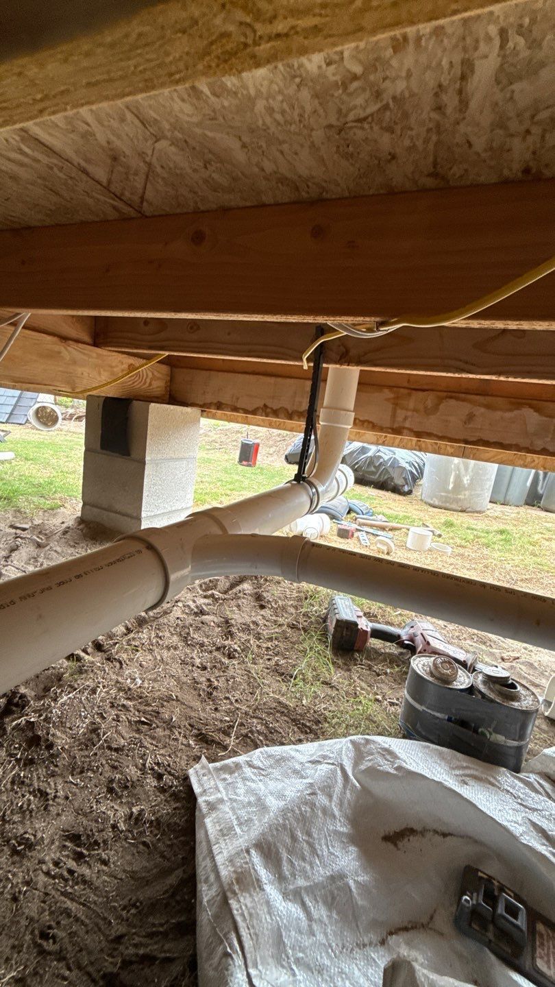 Under a wooden structure, a white pipe and cable run along beams above dirt and concrete blocks.
