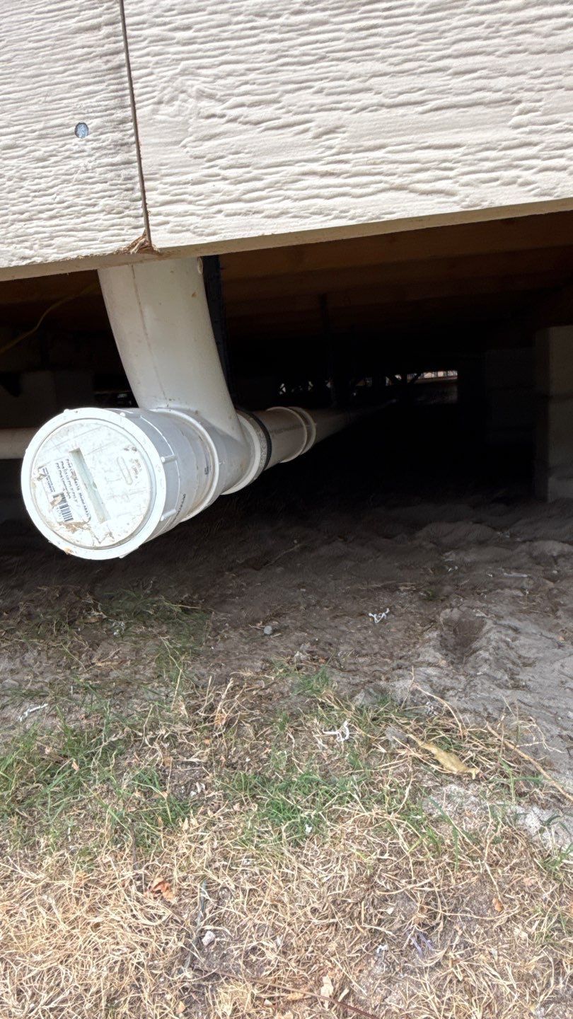 White pipe protruding under a house, extending into a crawlspace beneath beige siding.