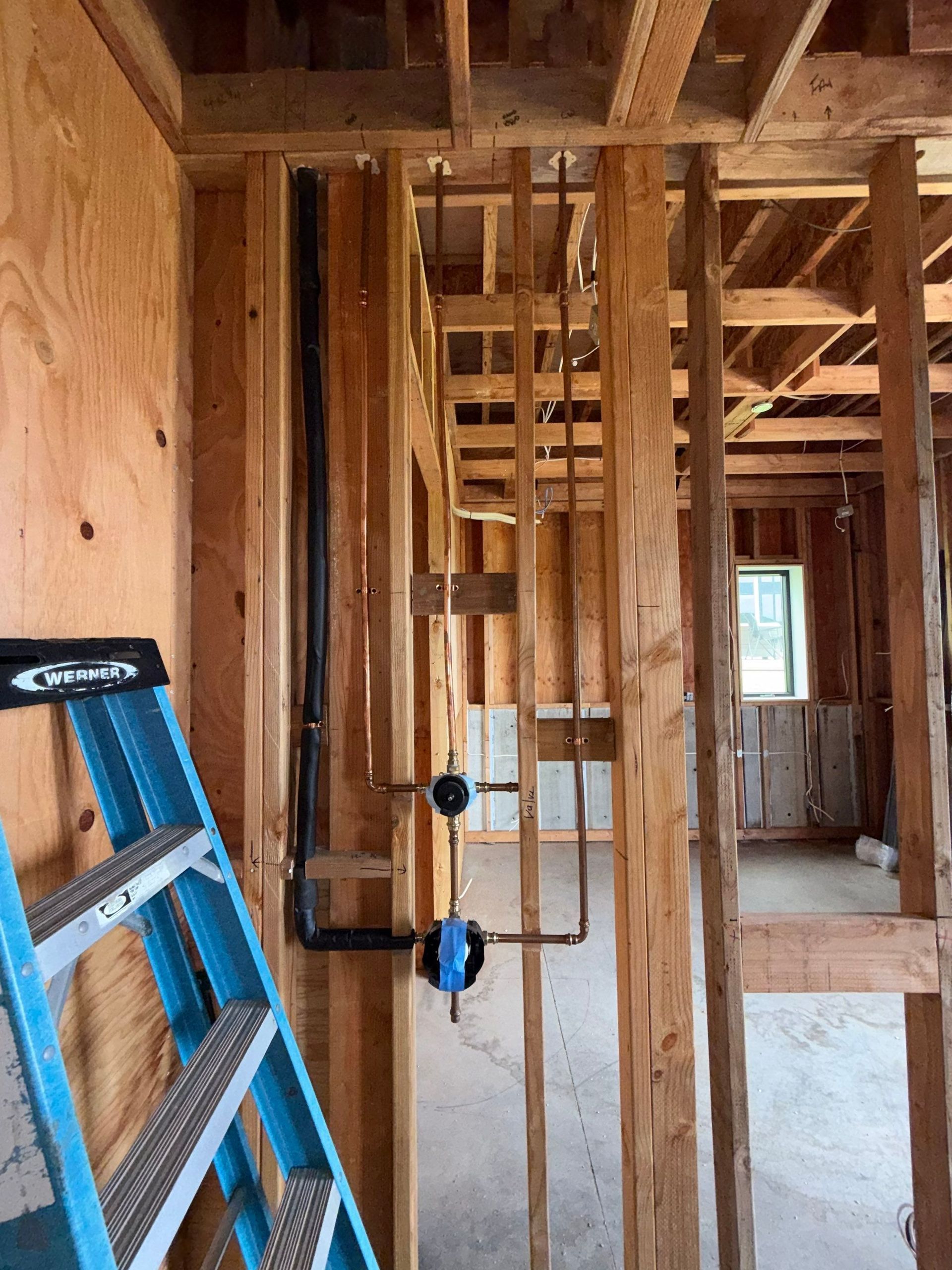 Interior of a house under construction with exposed wood framing, ladders, and a concrete floor.