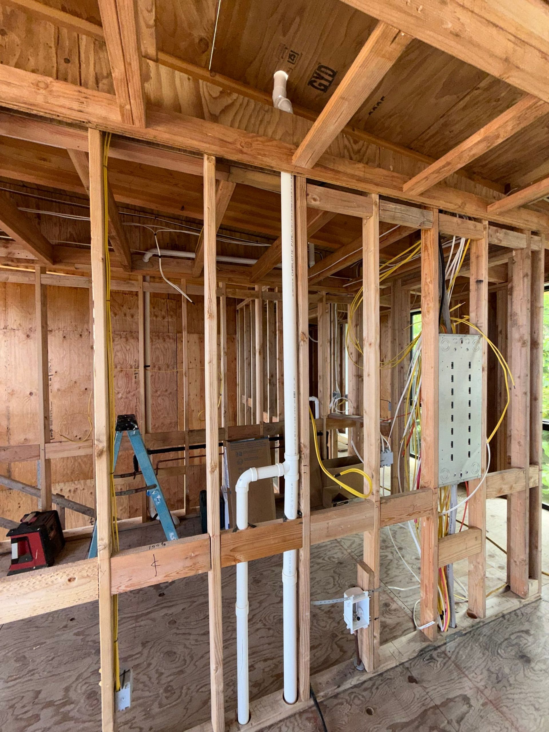 Framed interior wall under construction with exposed pipes, wiring, and a blue ladder in a unfinished room