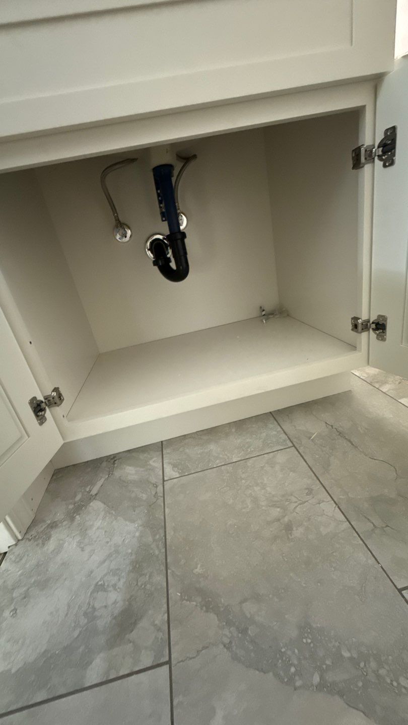 Open vanity cabinet under sink with exposed plumbing and gray tile floor