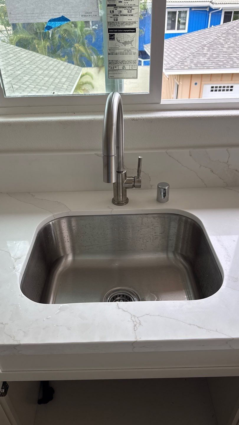 Stainless steel sink with tall faucet in a white countertop by a window with buildings outside