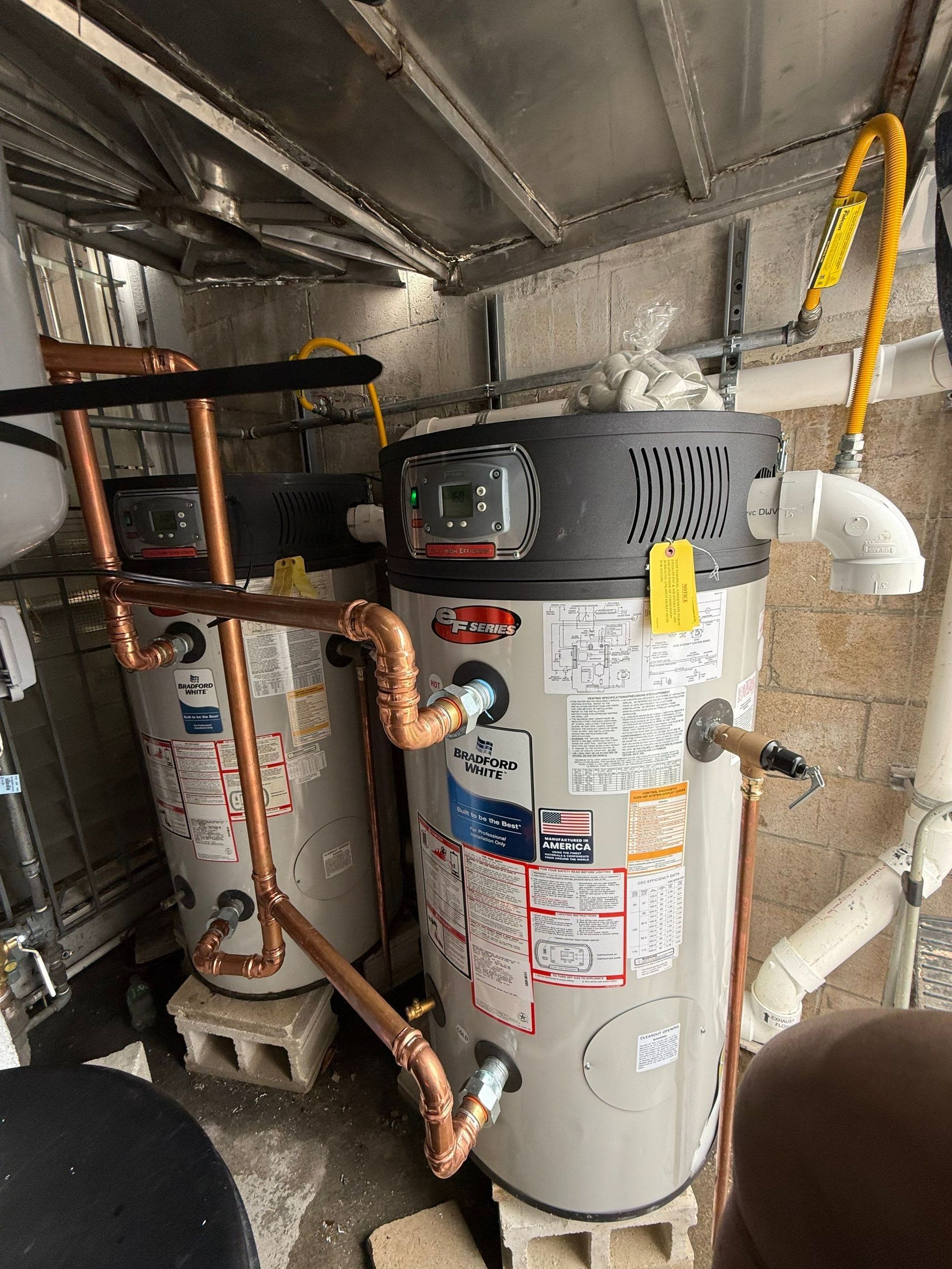 Two water heaters in a cramped utility closet with exposed pipes and insulated ducts.