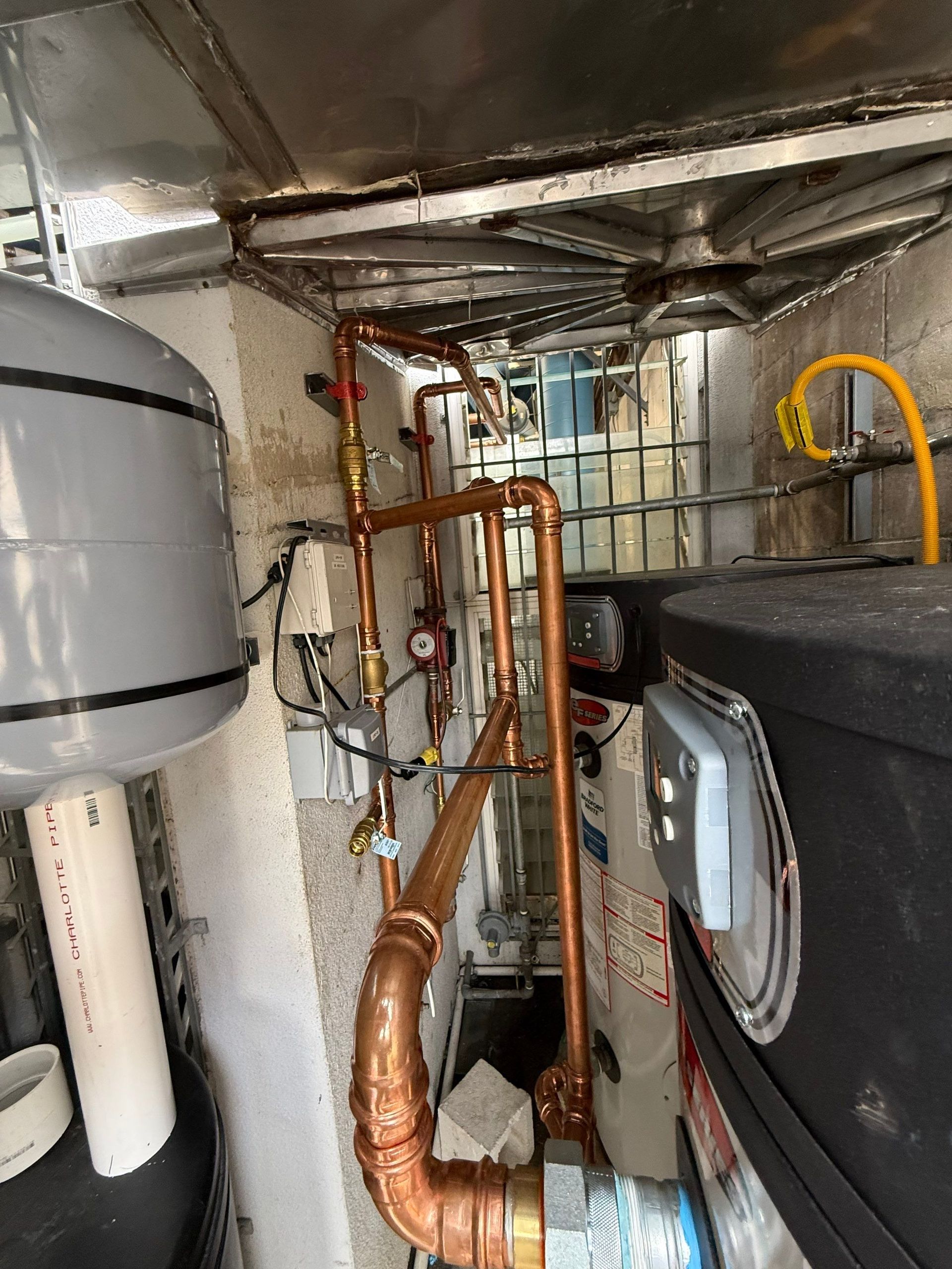 Narrow utility space with exposed copper pipes, white tank, and large black equipment beside insulated walls.