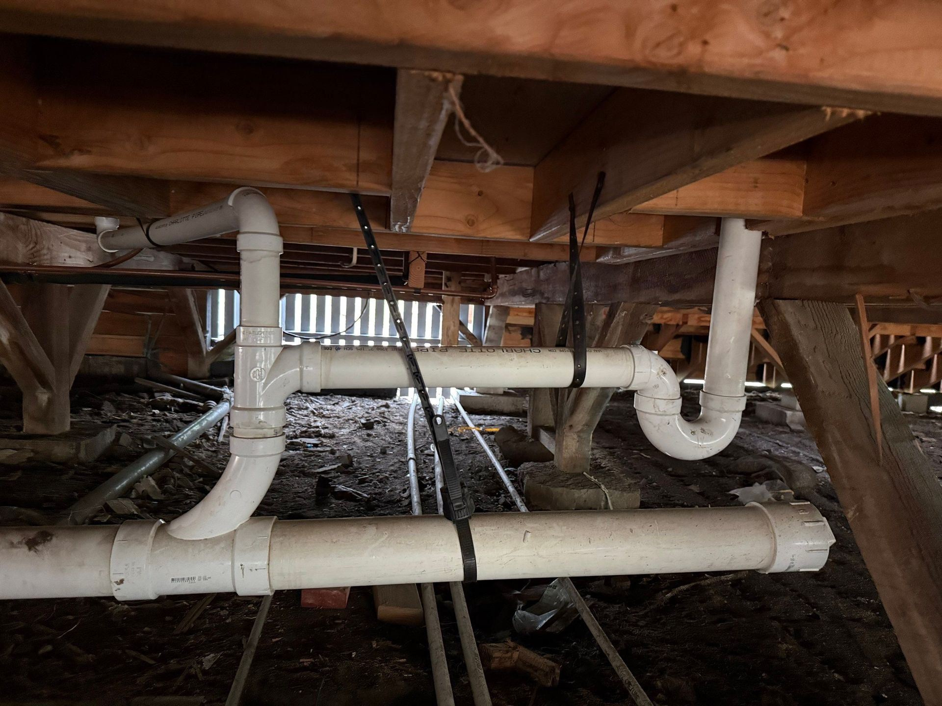 PVC plumbing pipes under a wooden floor crawlspace, with insulation and soil visible.