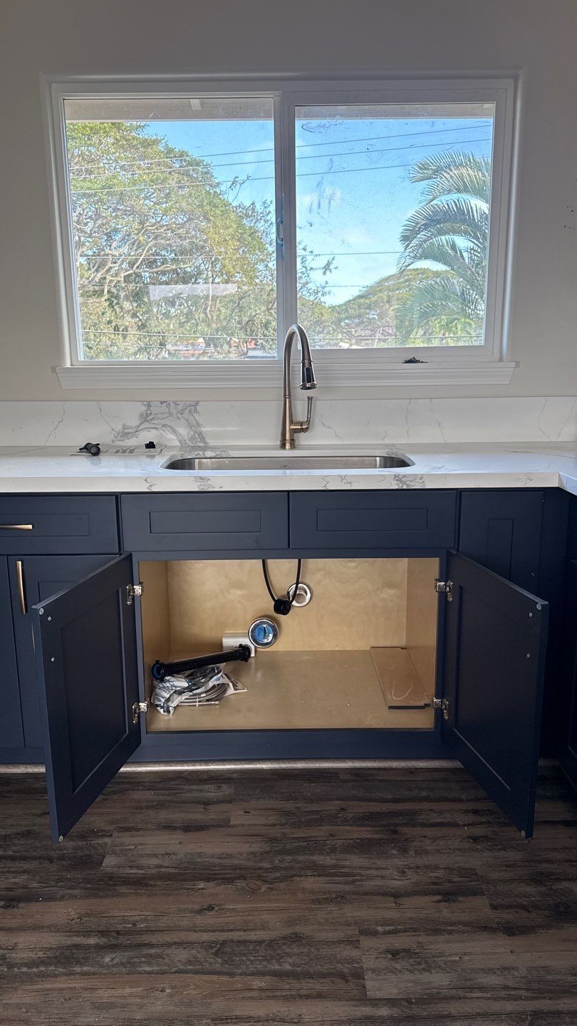 Kitchen sink with open dark cabinets beneath, showing exposed plumbing under a window.
