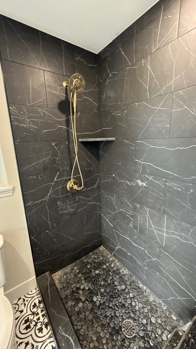 Black-tiled walk-in shower with brass rainfall showerhead and pebble floor in a modern bathroom
