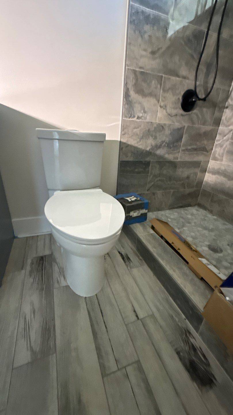 Bathroom with white toilet, gray tiled shower wall, and wood-look floor.