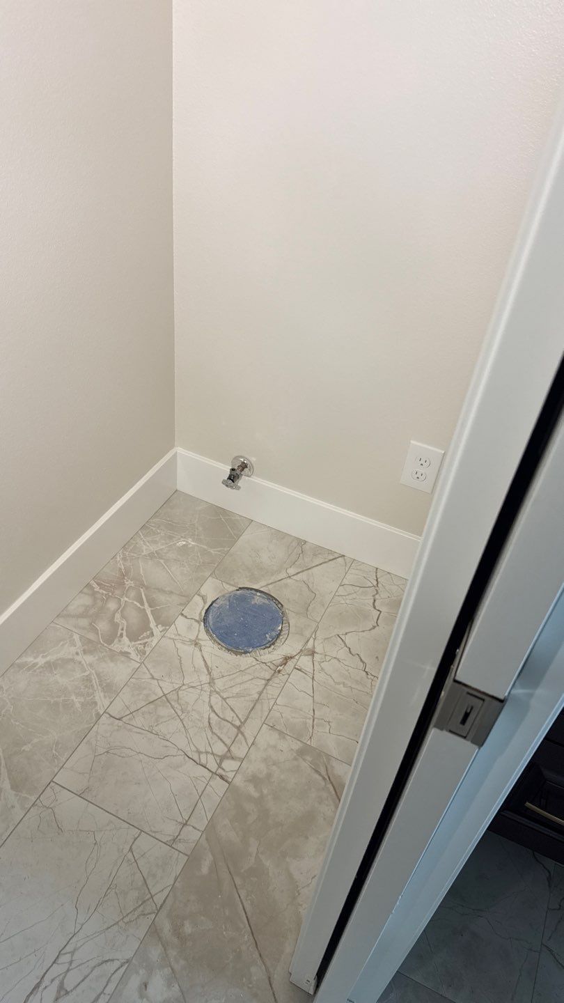 Small tiled laundry or utility nook with a floor drain, wall outlet, and plumbing hookup next to a doorway