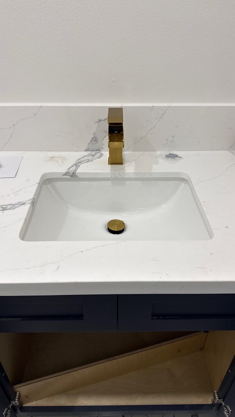 White bathroom sink with gold faucet and drain on a marble countertop; cabinet door open below.