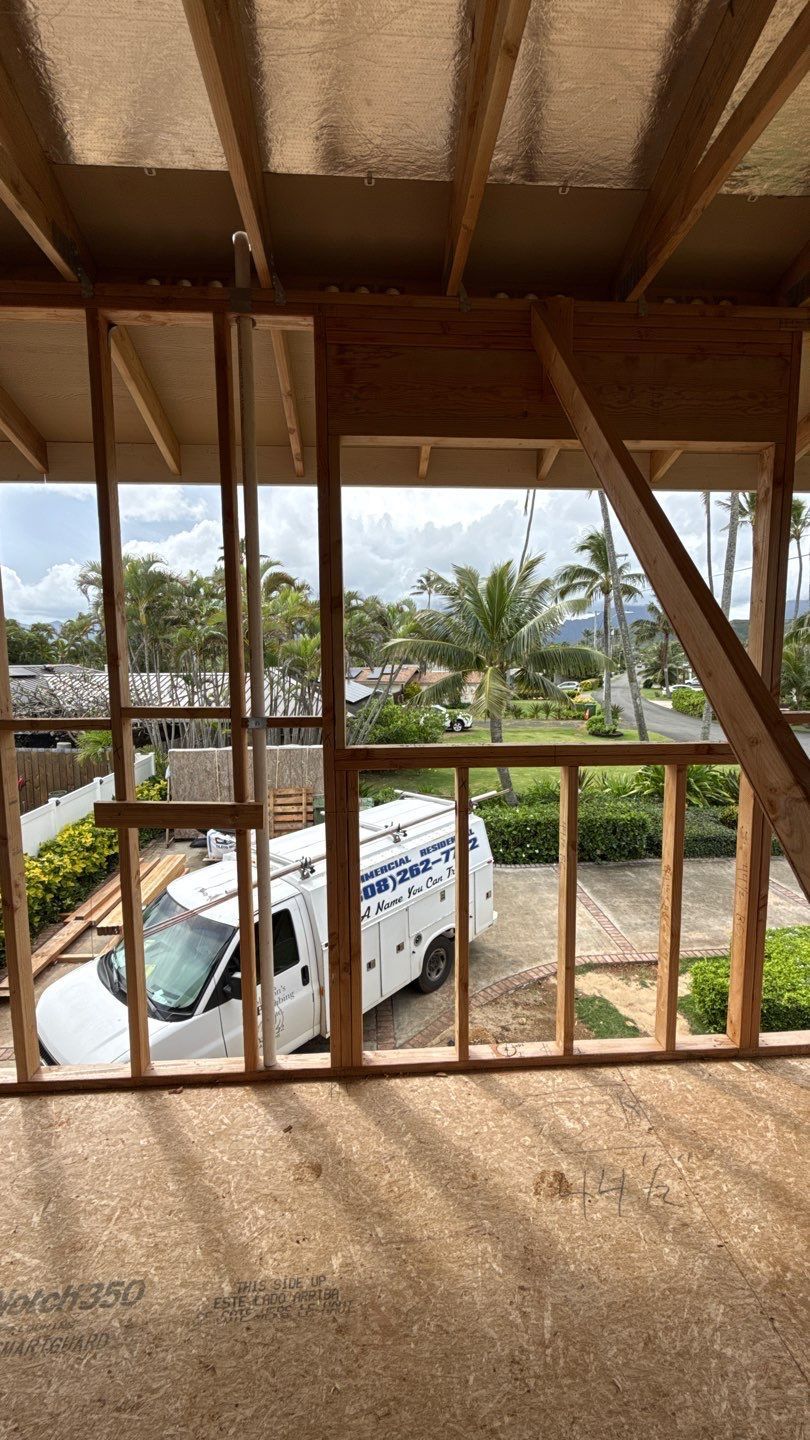 View from an unfinished room through wooden framing to a white van and palm trees outside.