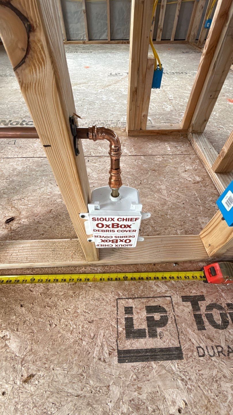Construction site floor with copper pipe and a small sign on plywood subfloor near a tape measure.