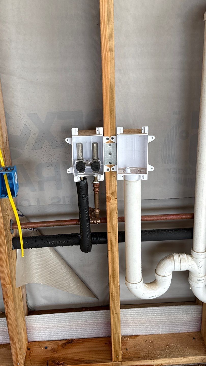 Small utility control box mounted on plywood beside white plumbing pipes in a wall cavity