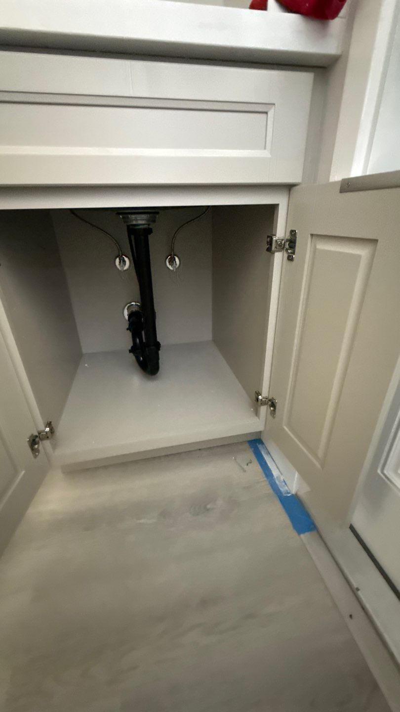 Open bathroom vanity cabinet with sink plumbing exposed and blue tape on the floor edge.