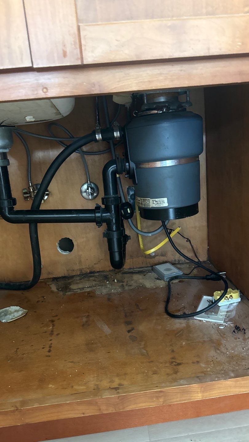 Under-sink plumbing with a gray disposal unit and exposed pipes in a wooden cabinet